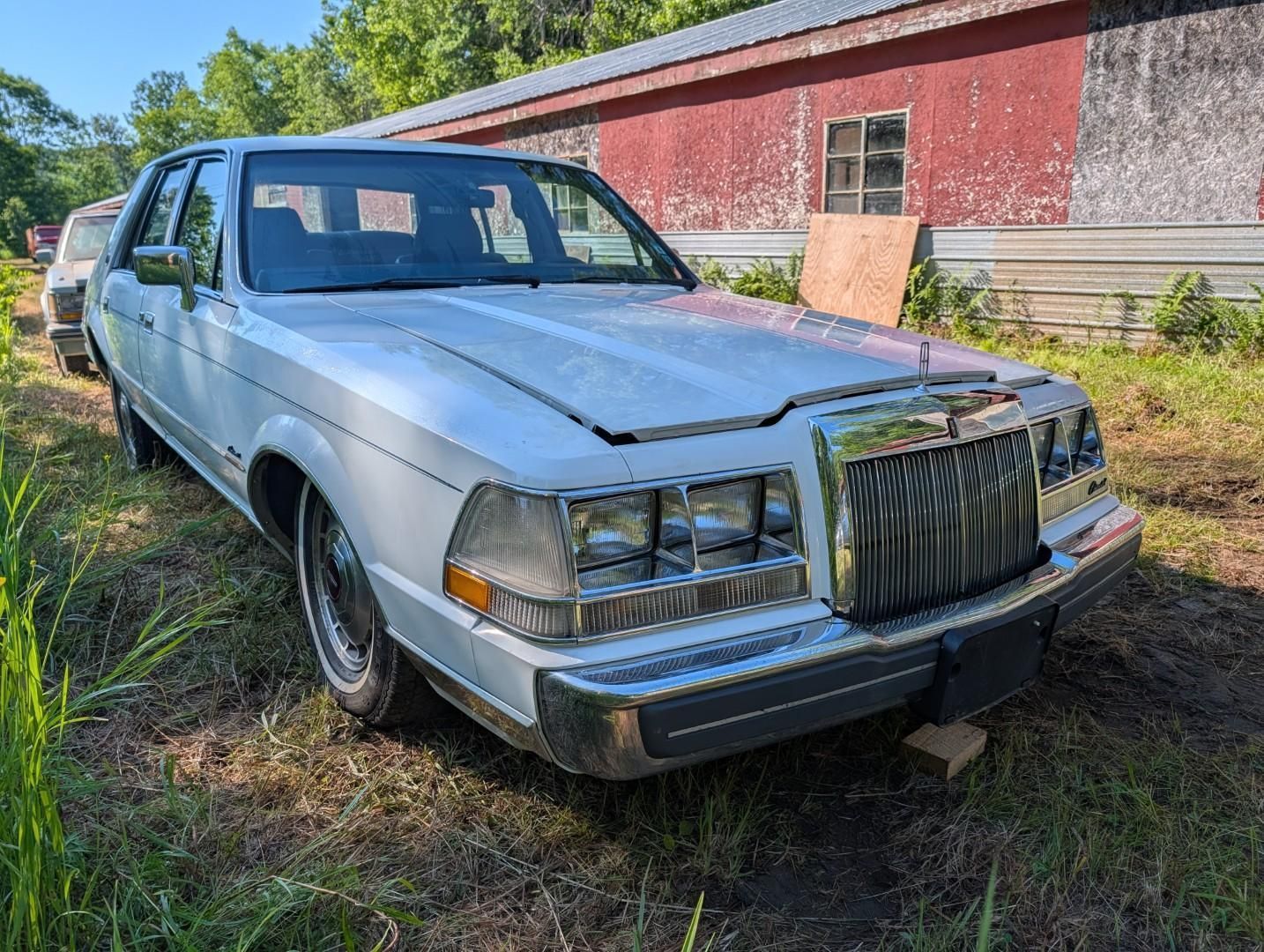 Old silver sedan parked on grass beside a red building, front view with chrome grille and hood ornament