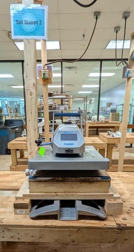 Blue heat press machine on a wooden table in a workshop or store, with shelves and tools in the background