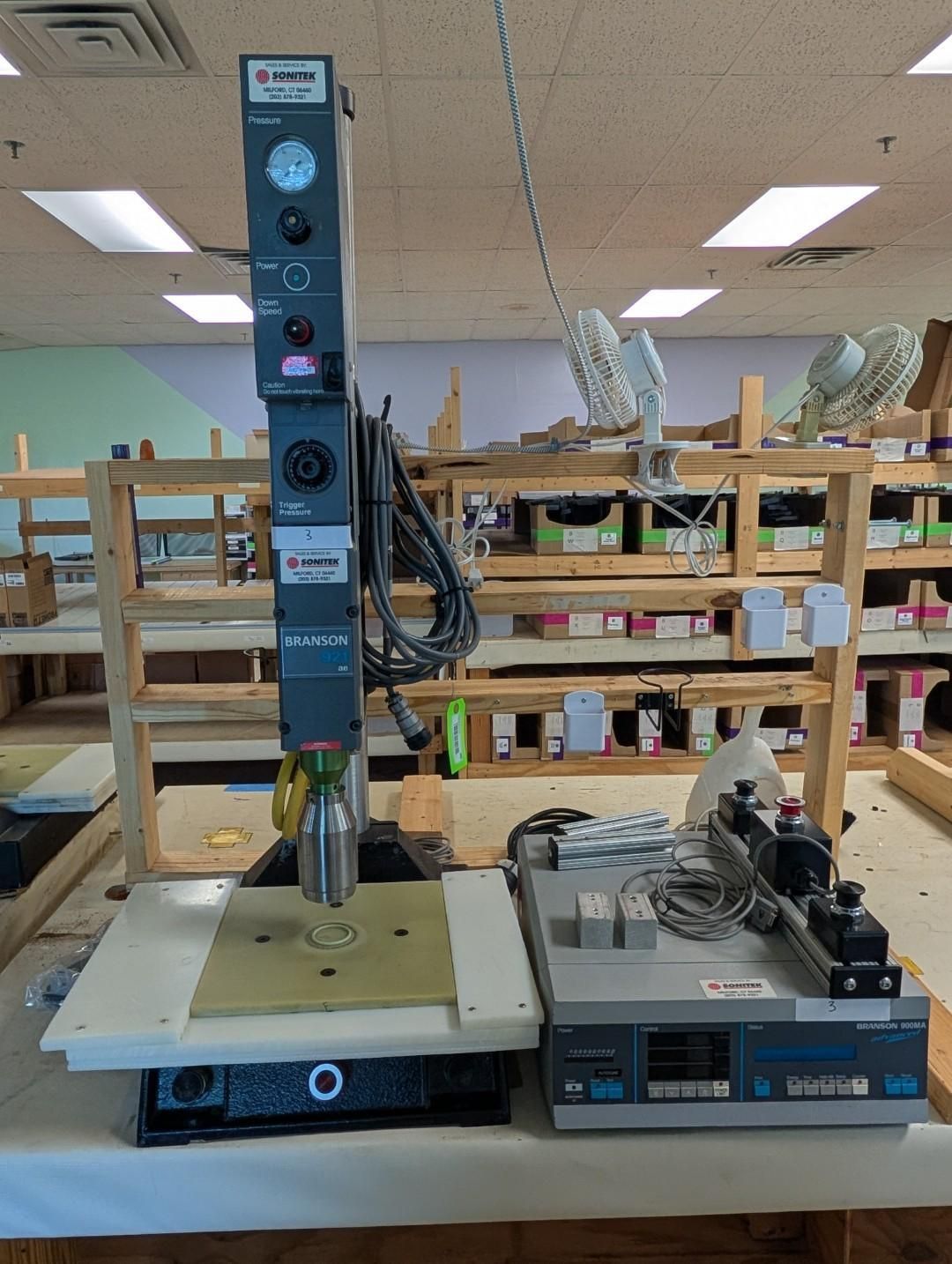 Industrial machine on a workbench in a workshop, with wooden shelving and equipment in the background.