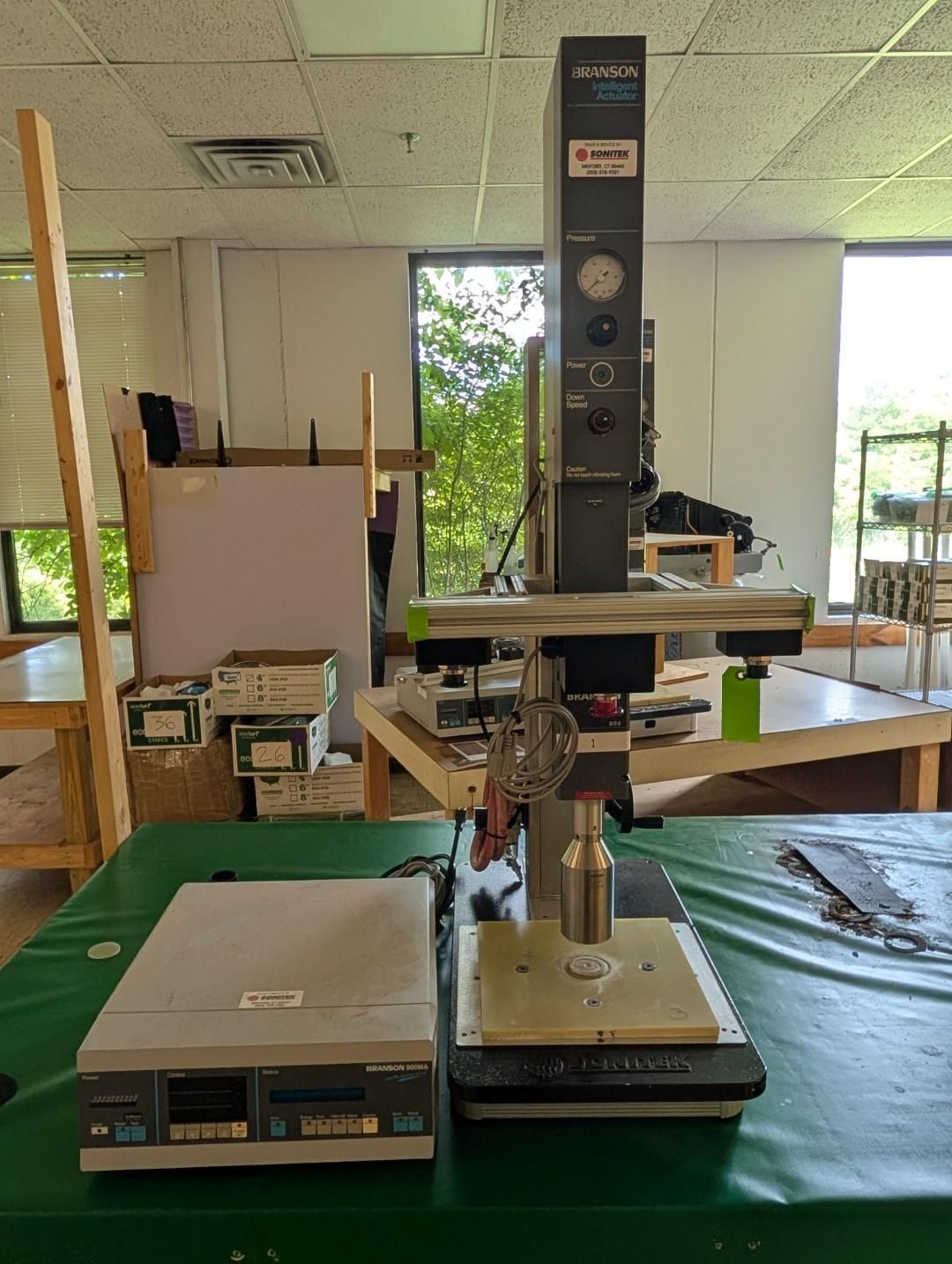 A lab workbench with a mounted testing machine, green cover, and a small control unit in a workshop.