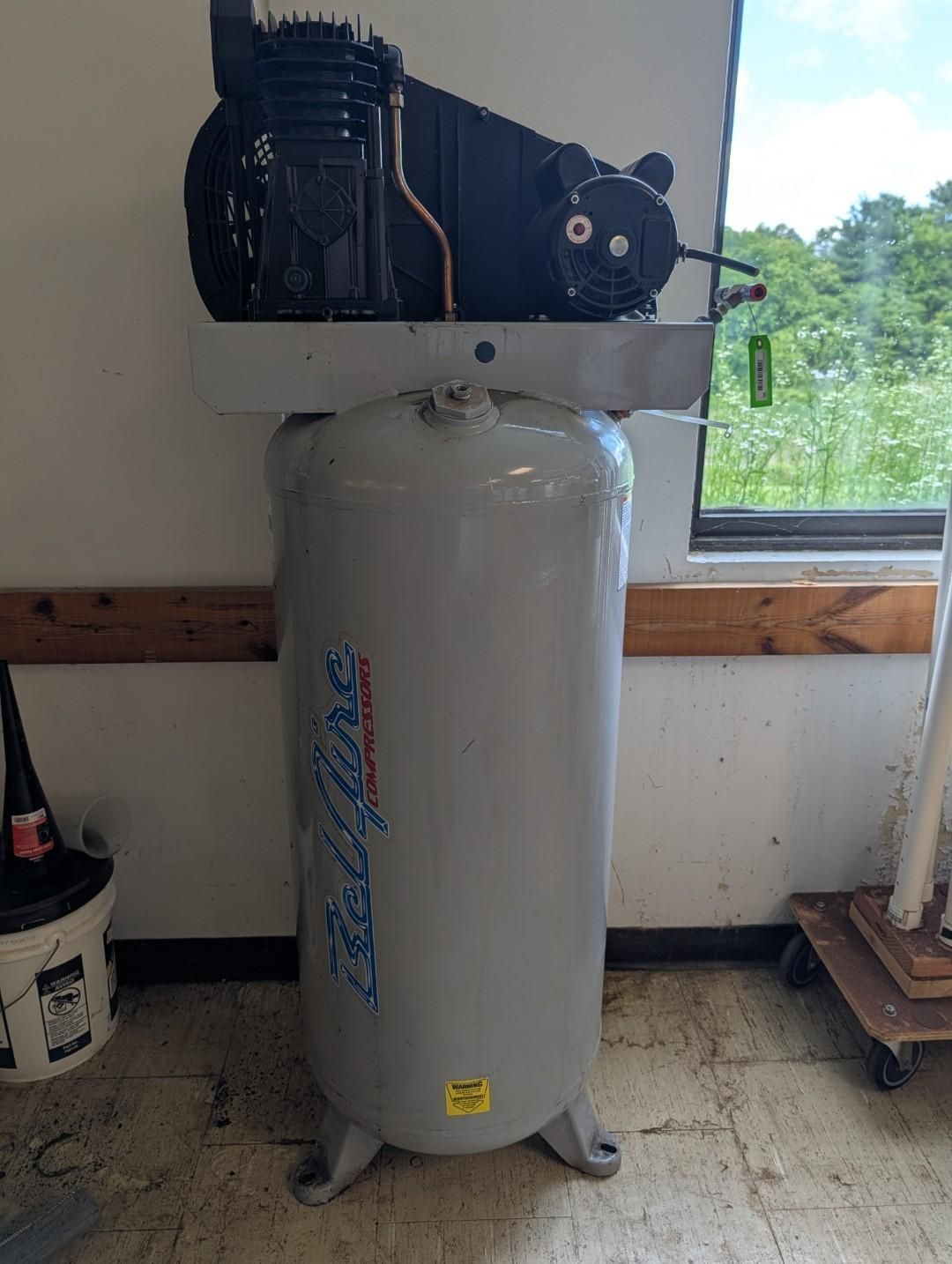 Gray air compressor in a workshop, with a black motor mounted on top near a window.