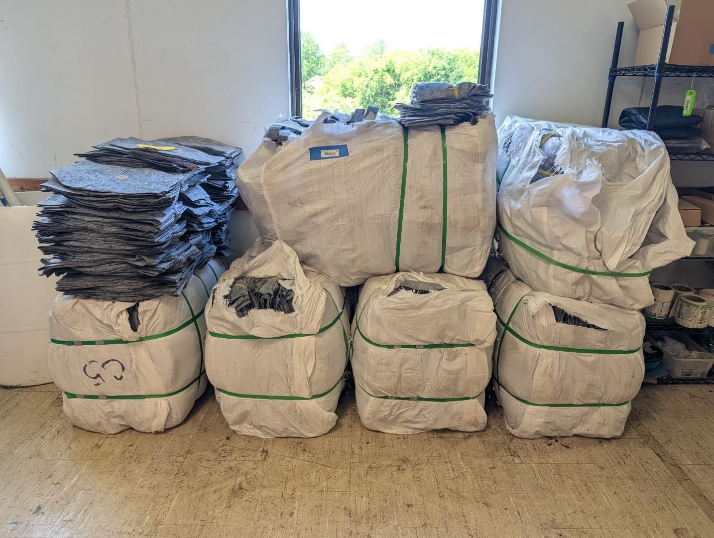 Stacked bundled materials in white sacks, with a pile of dark folded items on top, in a storage room.