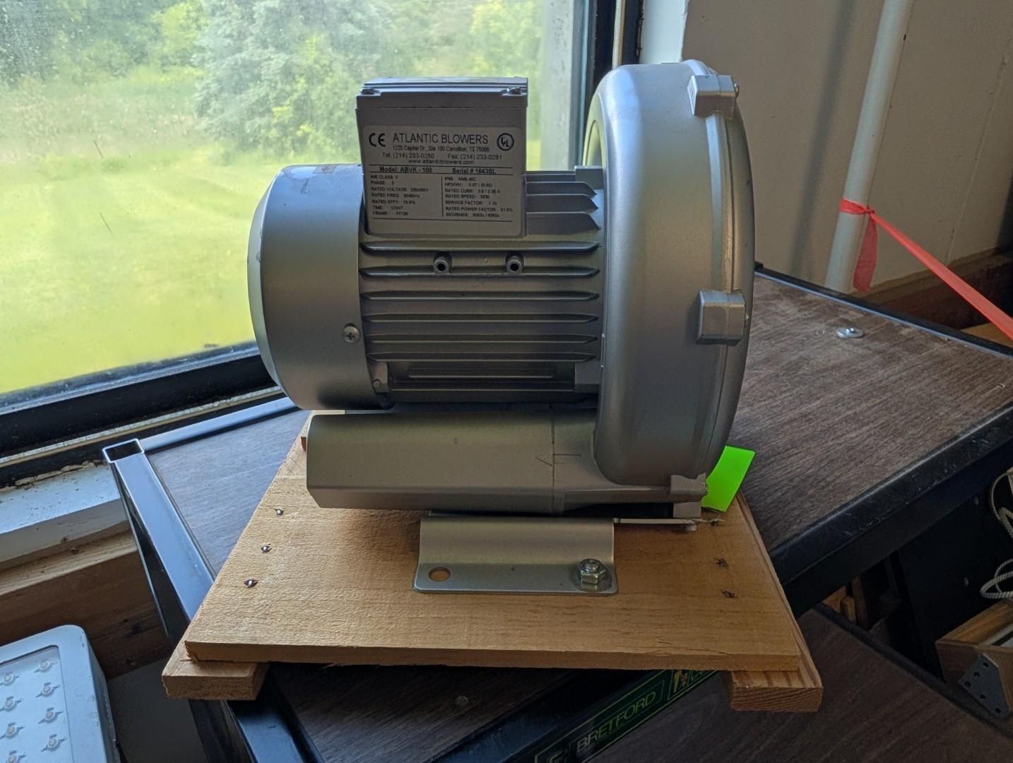 Electric motor mounted on a wooden board near a window, with a gray fan housing and green tag.