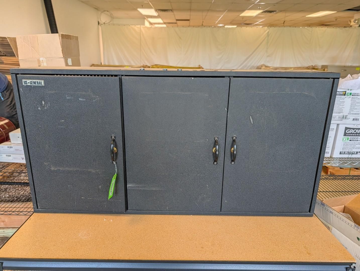 Gray wall cabinet with three doors above a tan countertop in a warehouse or workshop setting