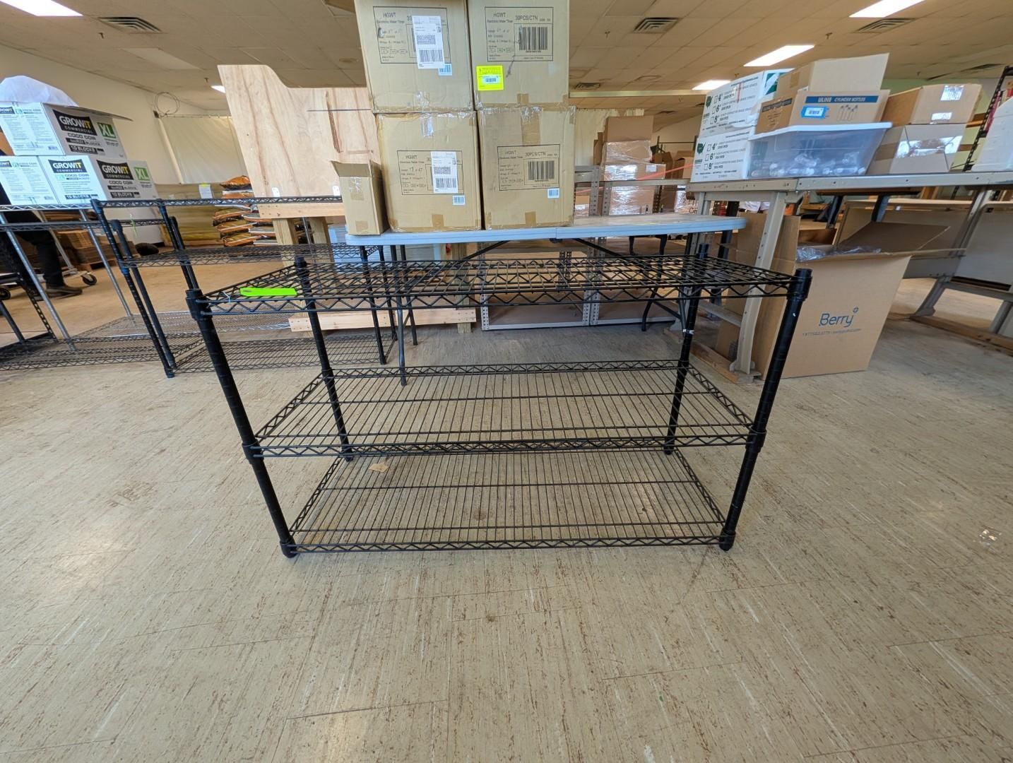 Black wire storage basket in a warehouse aisle with stacked cardboard boxes and tables behind it