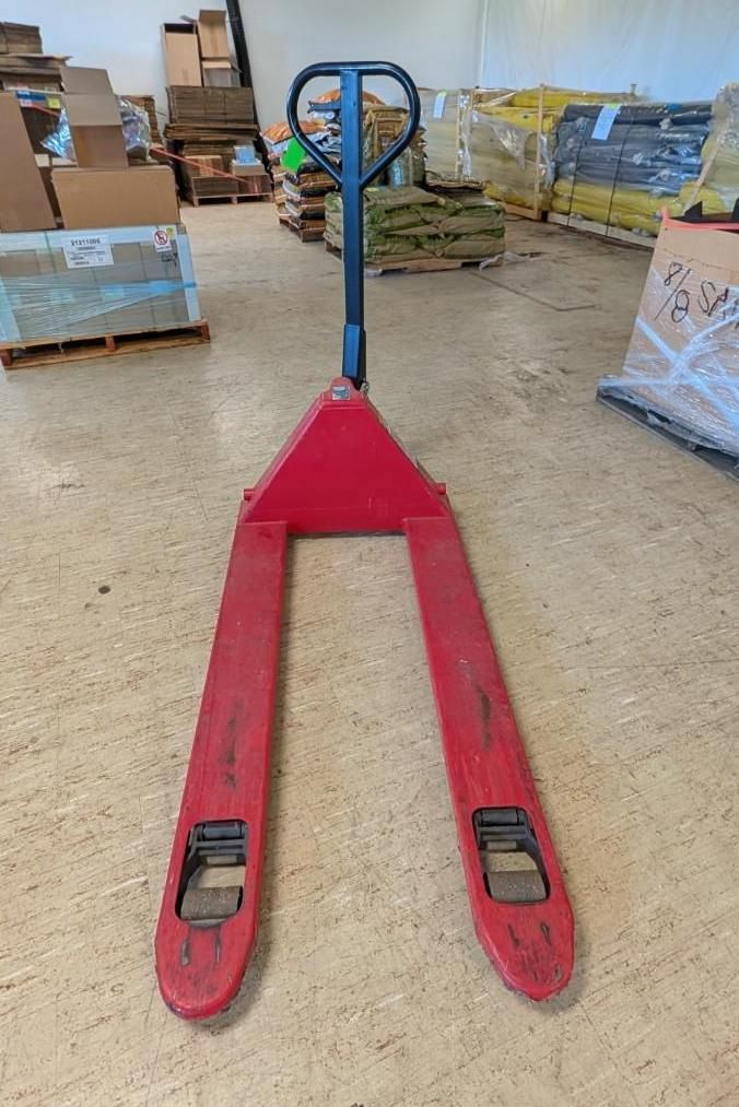 Red pallet jack in a warehouse, with blue handle and red forks on a concrete floor.