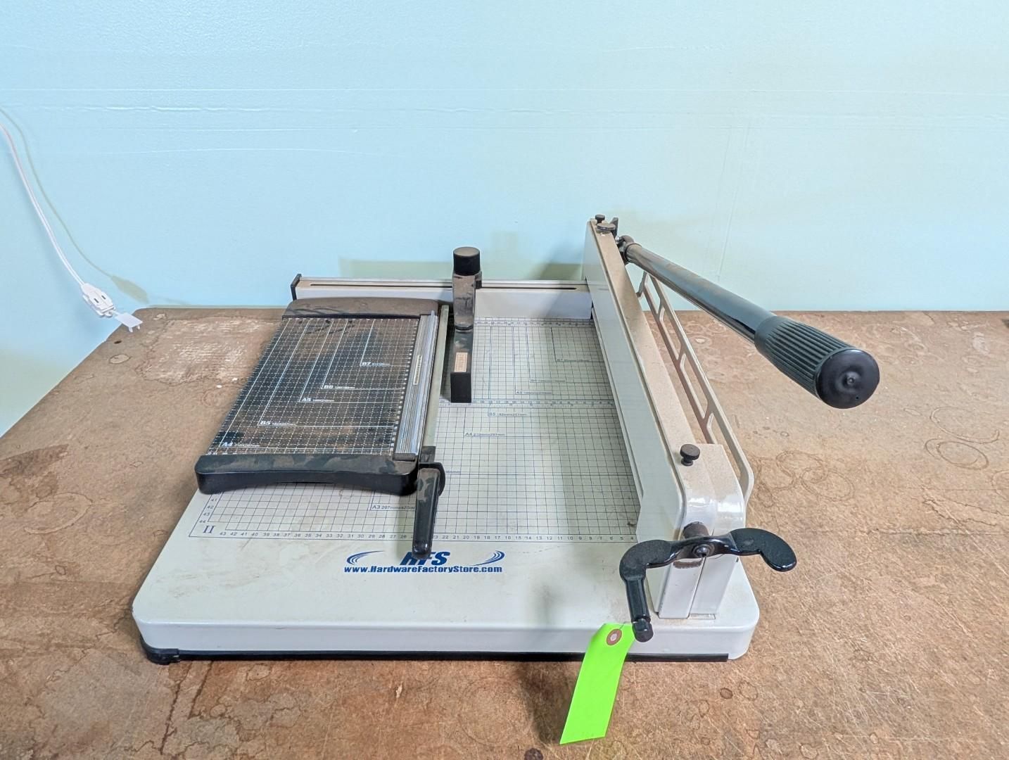 Paper cutter guillotine with metal arm, black handle, and green latch on a table against a blue wall