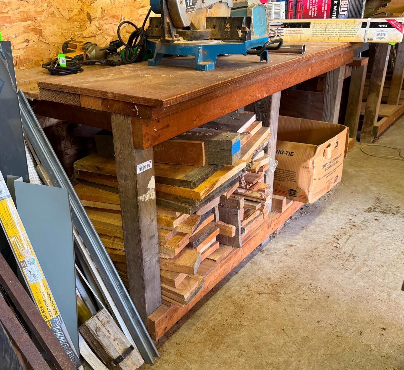 Wooden workbench in a workshop, with tools on top and lumber stored on shelves below.