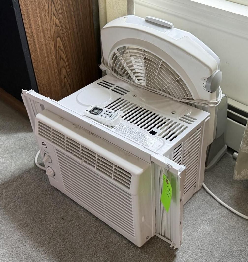 Two window air conditioners stacked on carpet beside a wall and window.