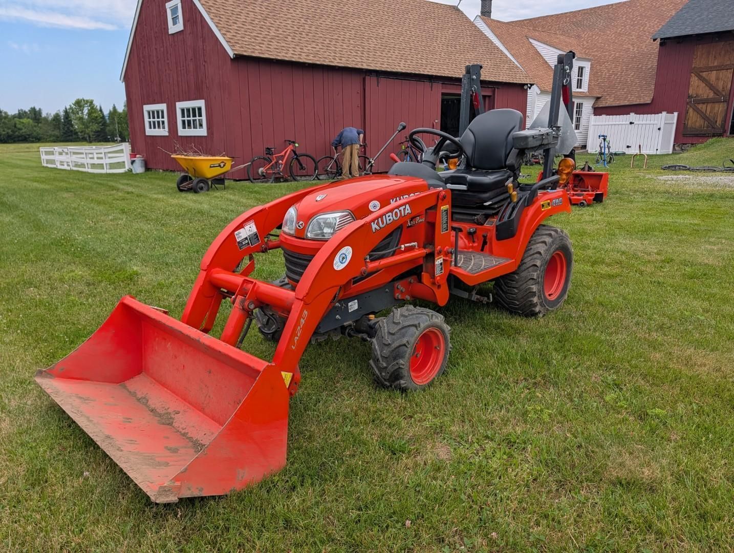 Orange tractor with front loader parked on grass beside a red barn and farm buildings