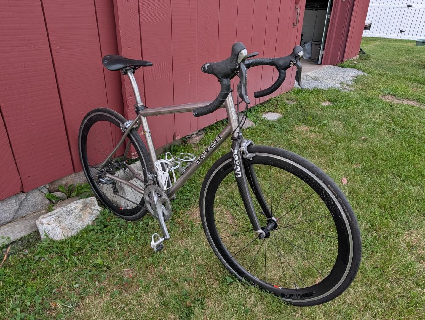 Black road bike leaning against a red wooden wall on grass.