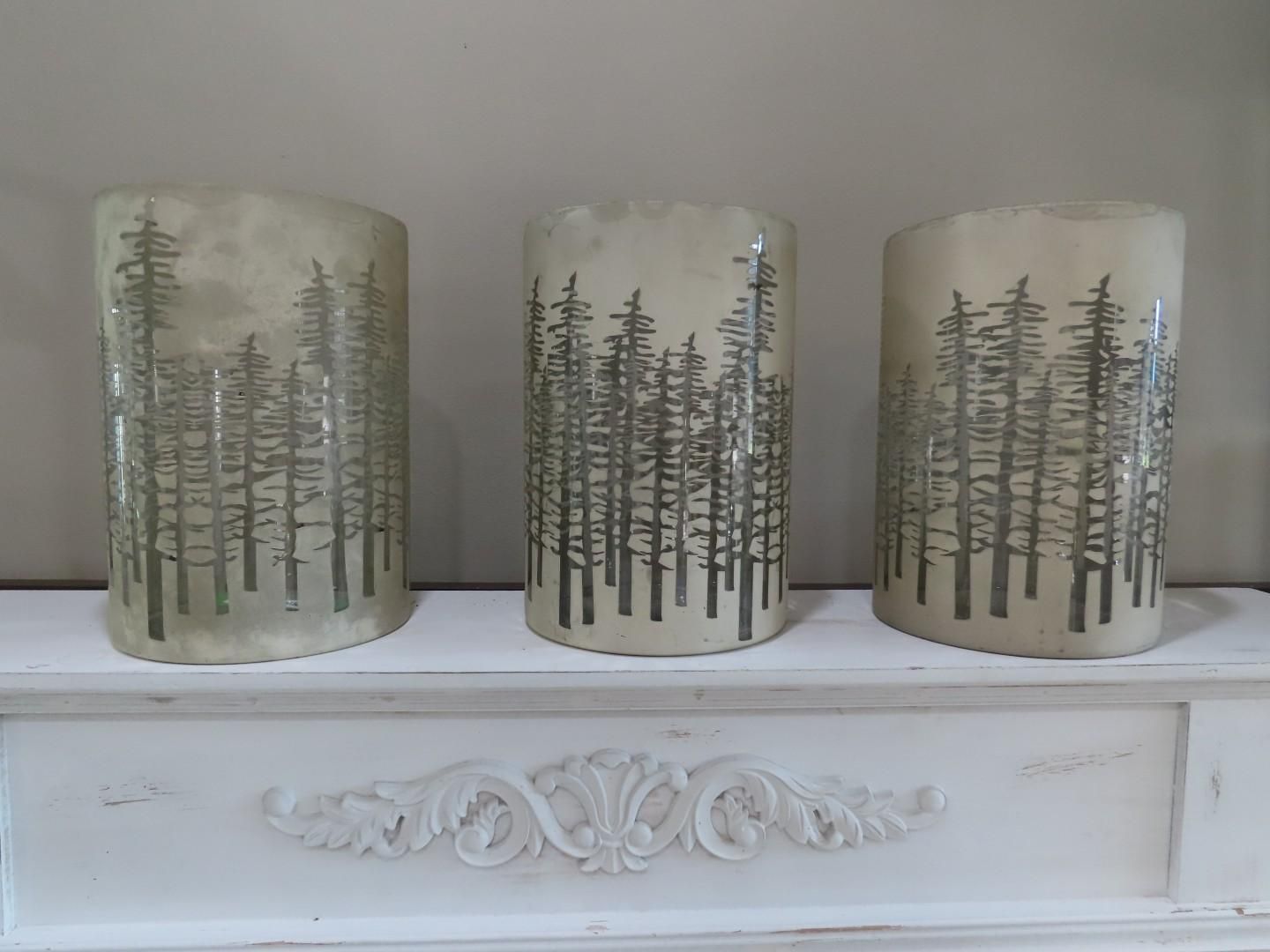 Three frosted glass candle holders with pine tree designs on a white mantel