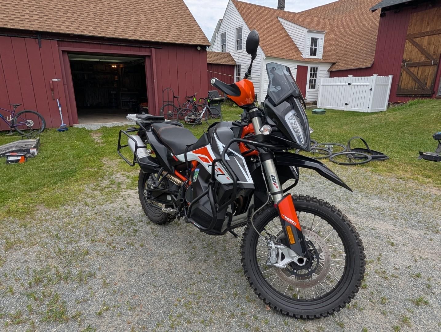 Black and orange off-road motorcycle parked on gravel beside a red barn and white buildings.