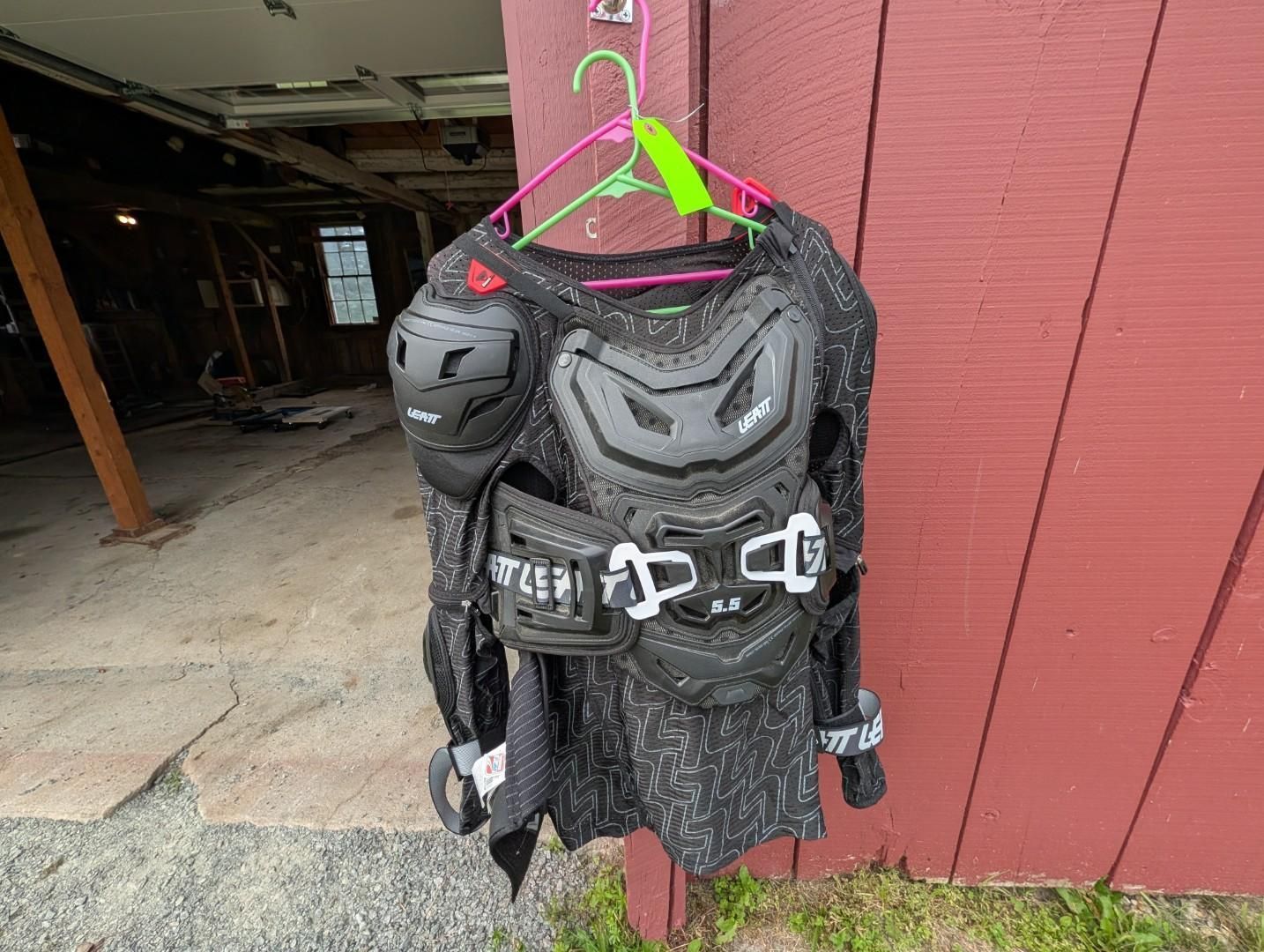 Black motocross protective gear hanging on a pink hanger beside a red barn wall.