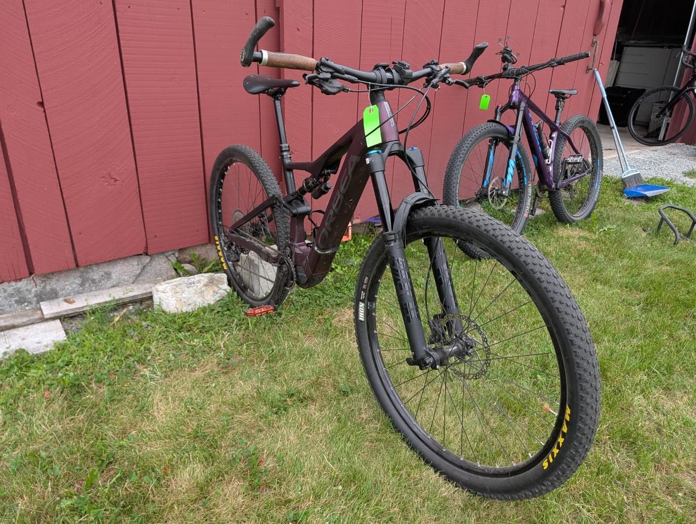 Two mountain bikes leaning against a red wooden wall on grass.
