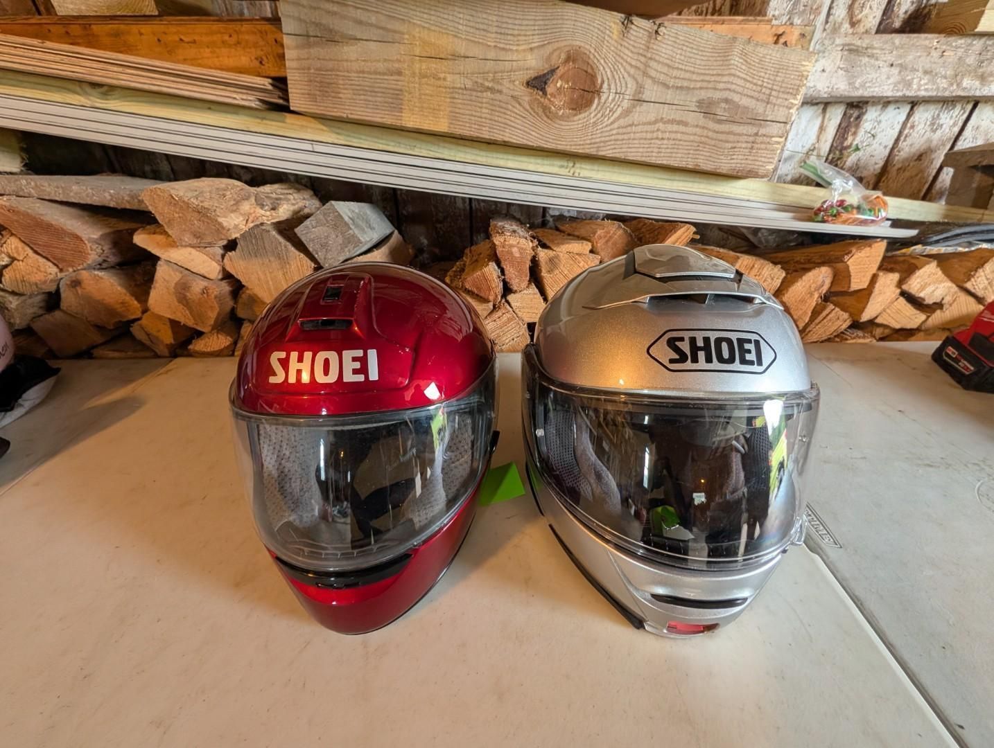 Two SHOEI motorcycle helmets, red and silver, sitting on a floor in a wood storage area