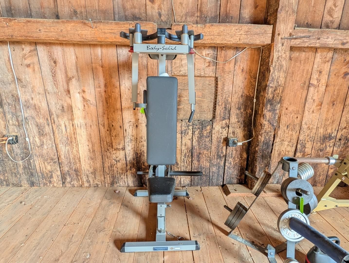 Home gym bench press machine with padded seat and barbell rack on a wooden floor.