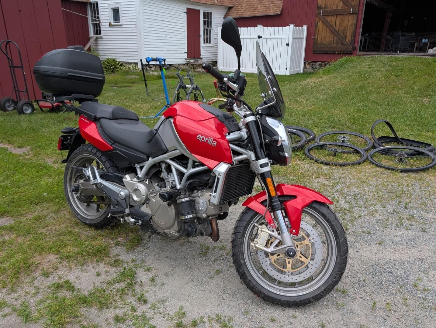 Red motorcycle parked on grass in a yard beside a house and bicycles.