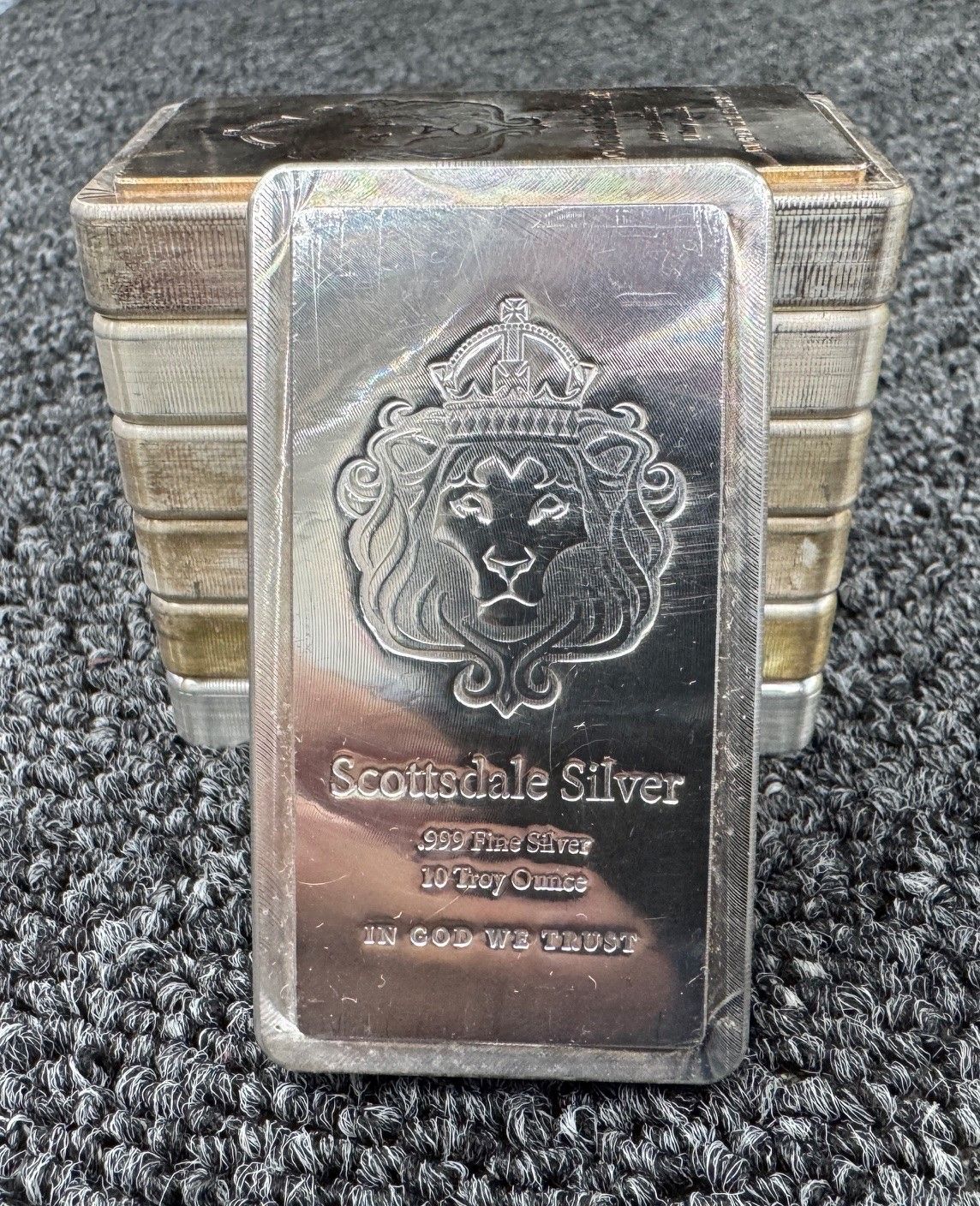 Stack of silver bars, front bar labeled “Scottsdale Silver” with lion emblem, on gray carpet.