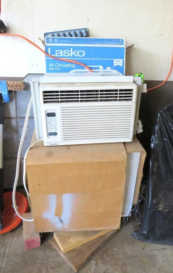 White air conditioner on a cardboard box, with a blue Lasco box on top, against a wall.