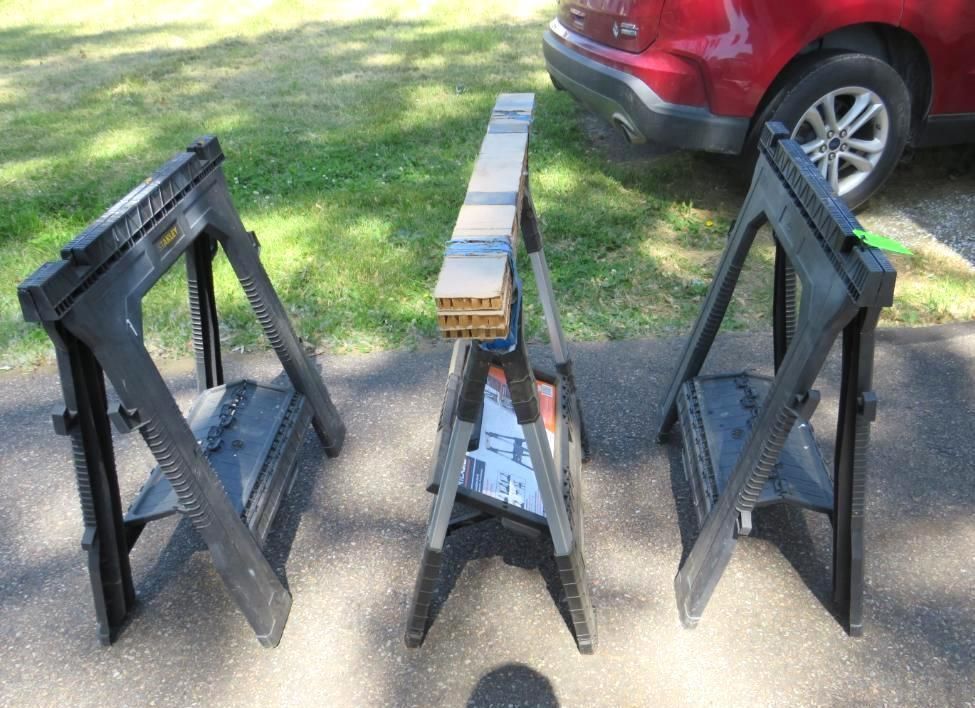 Three black sawhorses set up on asphalt with a board on top.