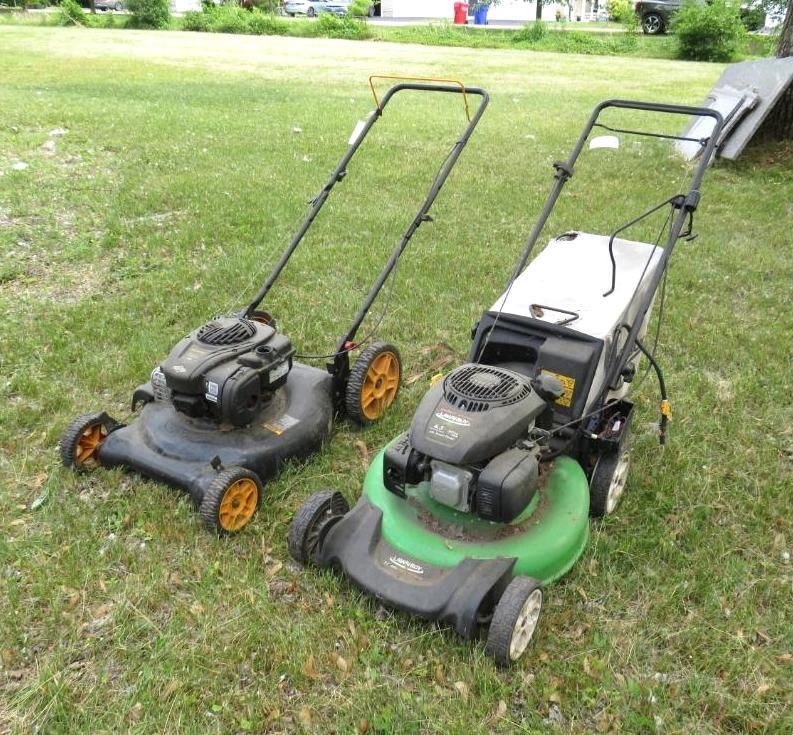 Two gas-powered lawn mowers sit on a grassy lawn. One has yellow wheels, the other white.