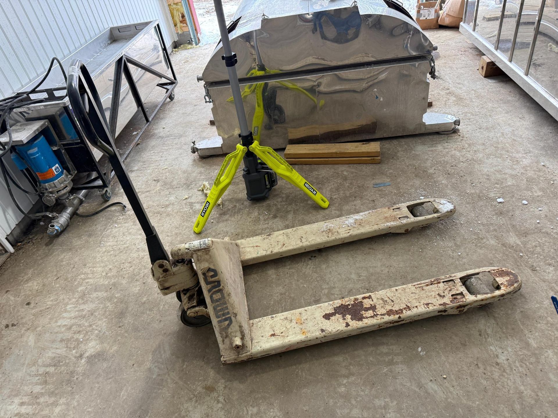 White pallet jack on a concrete floor with large metal object and work light in background.
