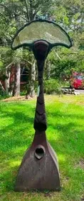 Sculpture on a grassy lawn. The dark, abstract art has a fan-shaped top, a long neck, and a triangular base.