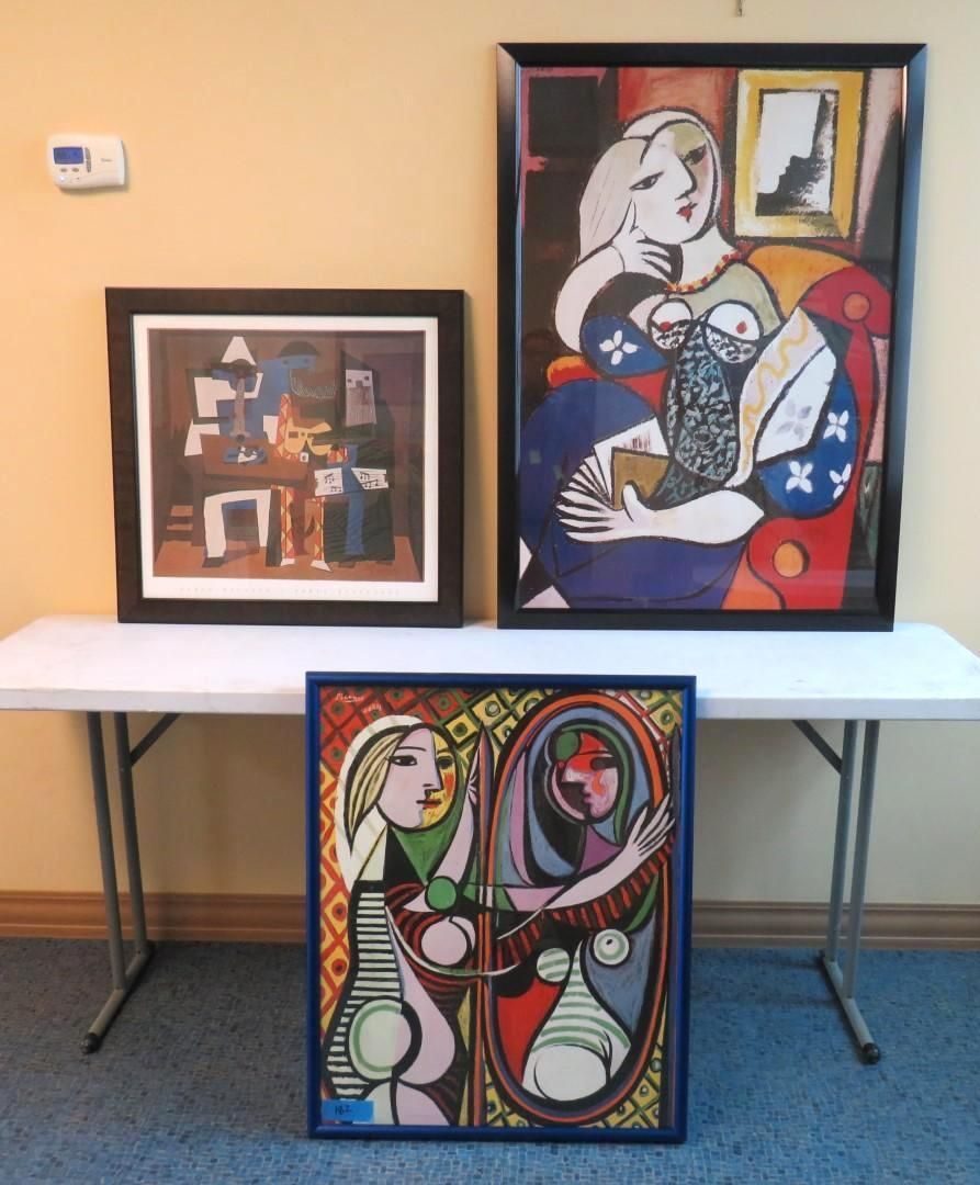 Three framed paintings on a white table. One painting shows an abstract interior, two show Picasso-esque portraits.