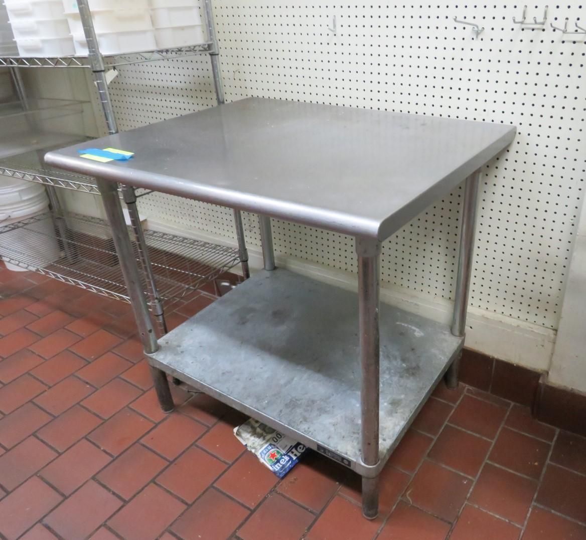 Stainless steel work table with a lower shelf, on red tile floor, beside pegboard wall.