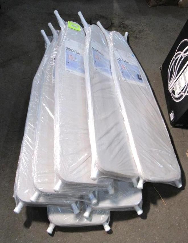 Stack of wrapped ironing boards, light grey, stacked on a concrete floor.