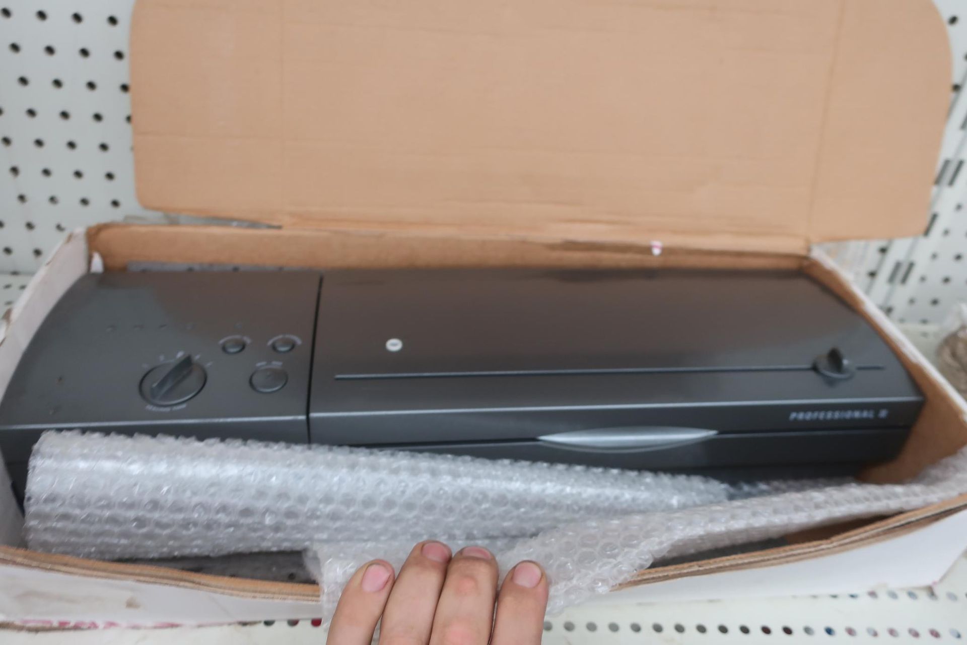 A dark gray laminator in its cardboard box, protected by bubble wrap, on a store shelf.