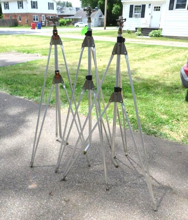 Three tall silver tripods with sprinkler heads set on a driveway.