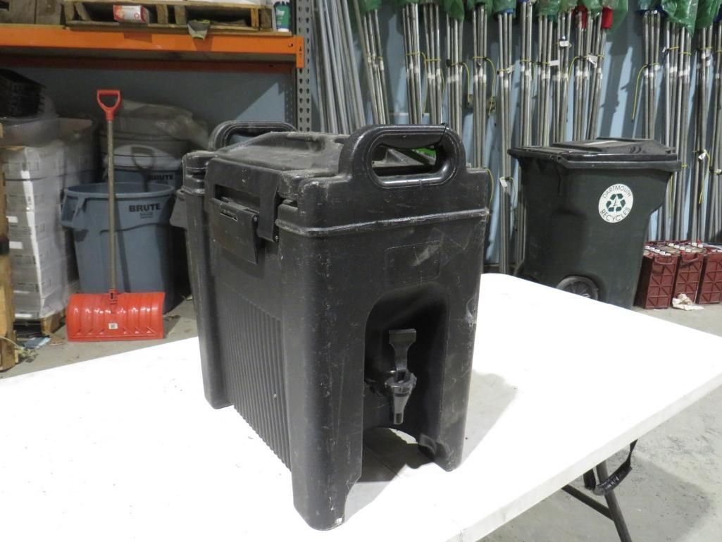 Black insulated beverage container on a white table, with spigot, handle, and latches. In a storage setting.