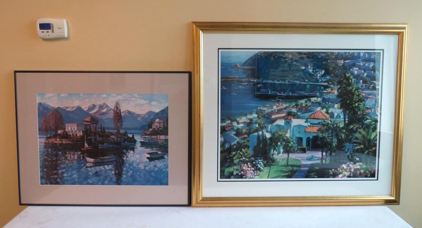 Two framed landscape paintings on a wall: lake scene with mountains, and a coastal town view.