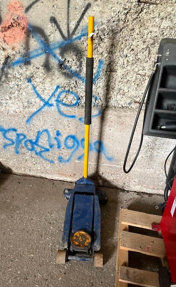 Blue and yellow floor jack with black handle in a garage, next to a pallet.