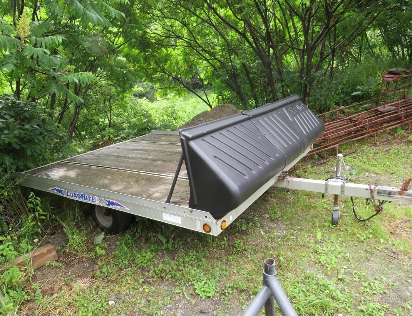A snowmobile trailer with a black rear guard parked on a grassy slope next to trees.