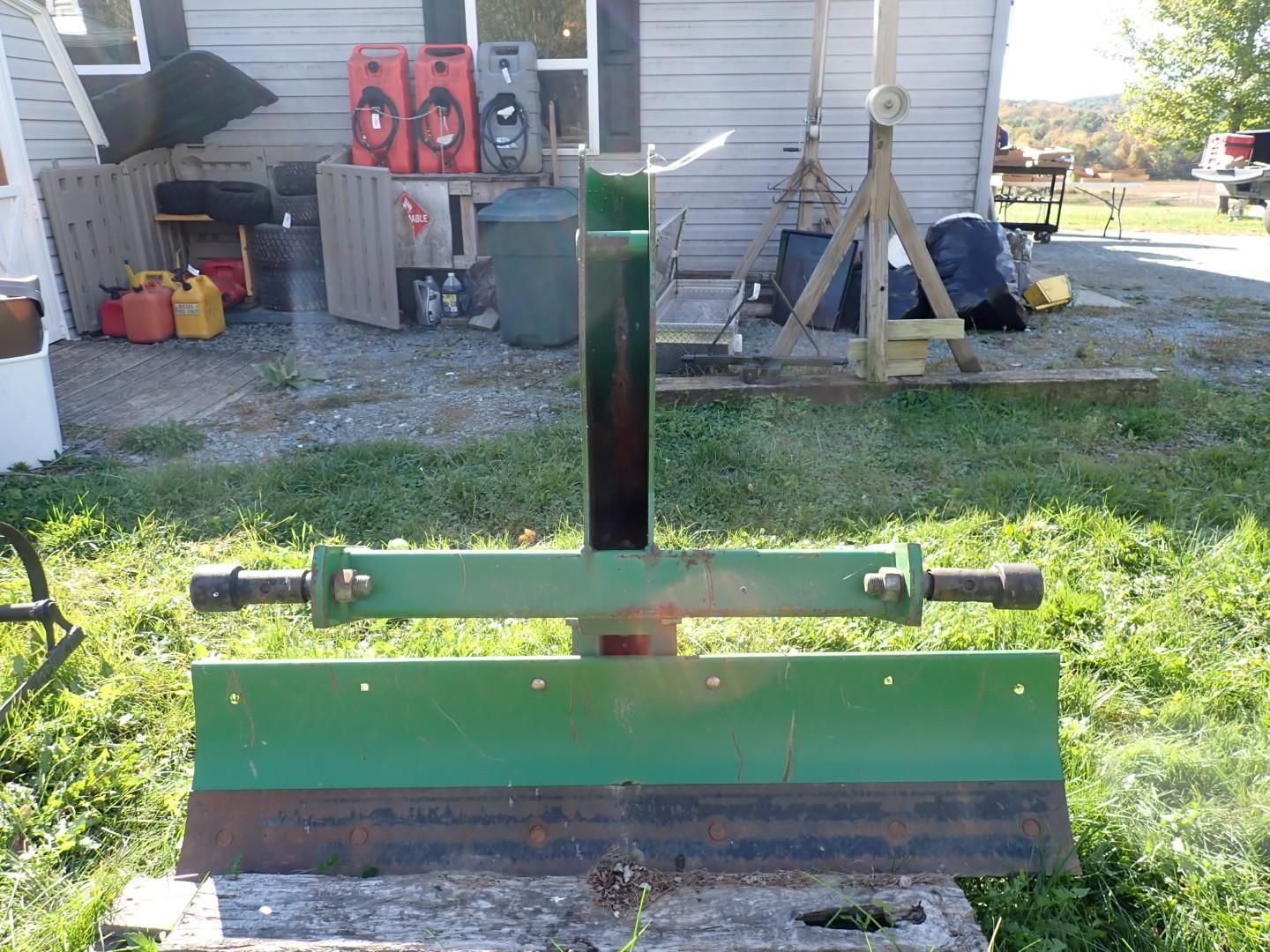 Green snowplow blade attachment standing in a gravel yard beside a house, with equipment and tools in the background.