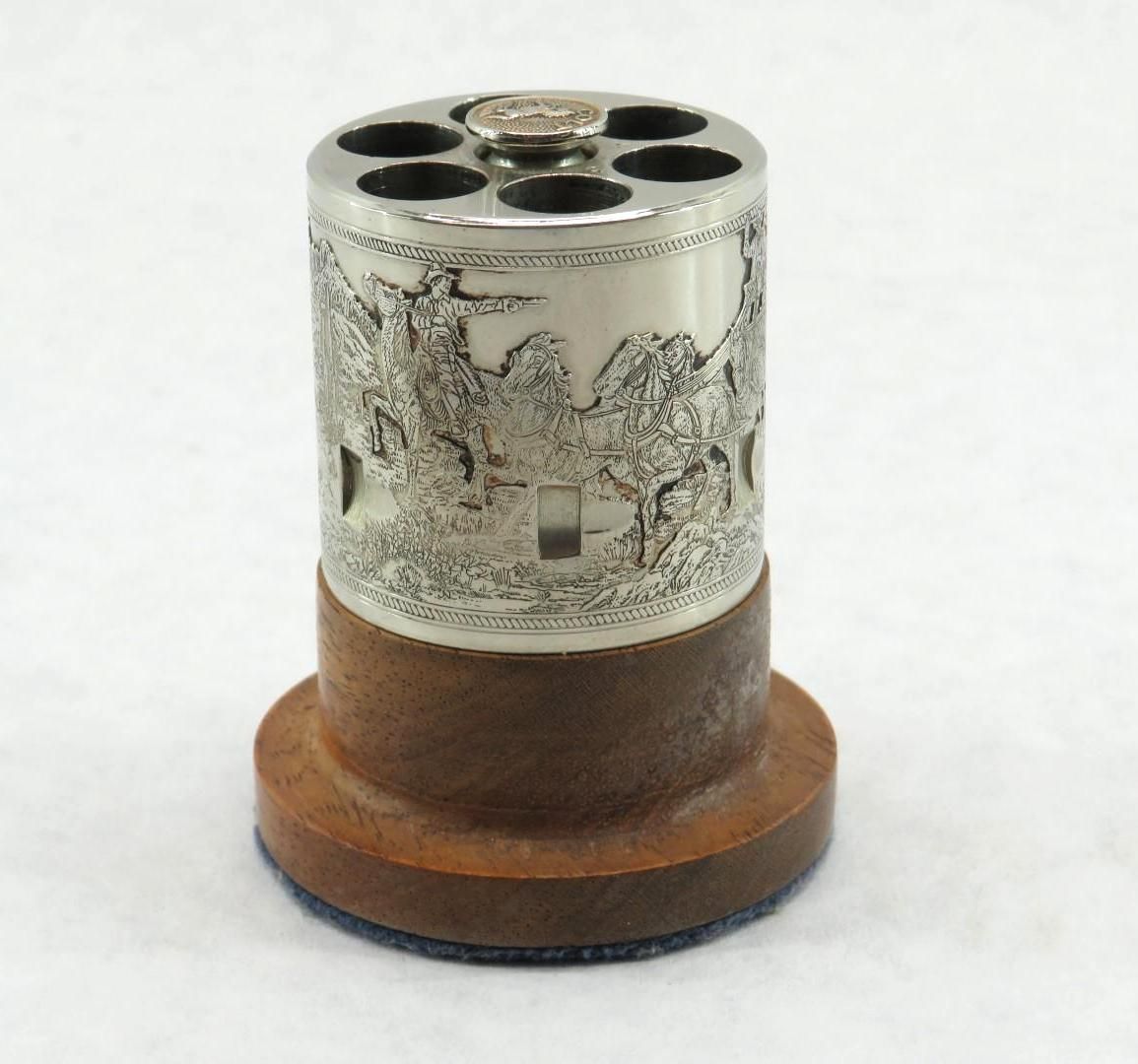 Silver firearm chamber, cylindrical pencil holder with engraved landscape scene on a wooden base.