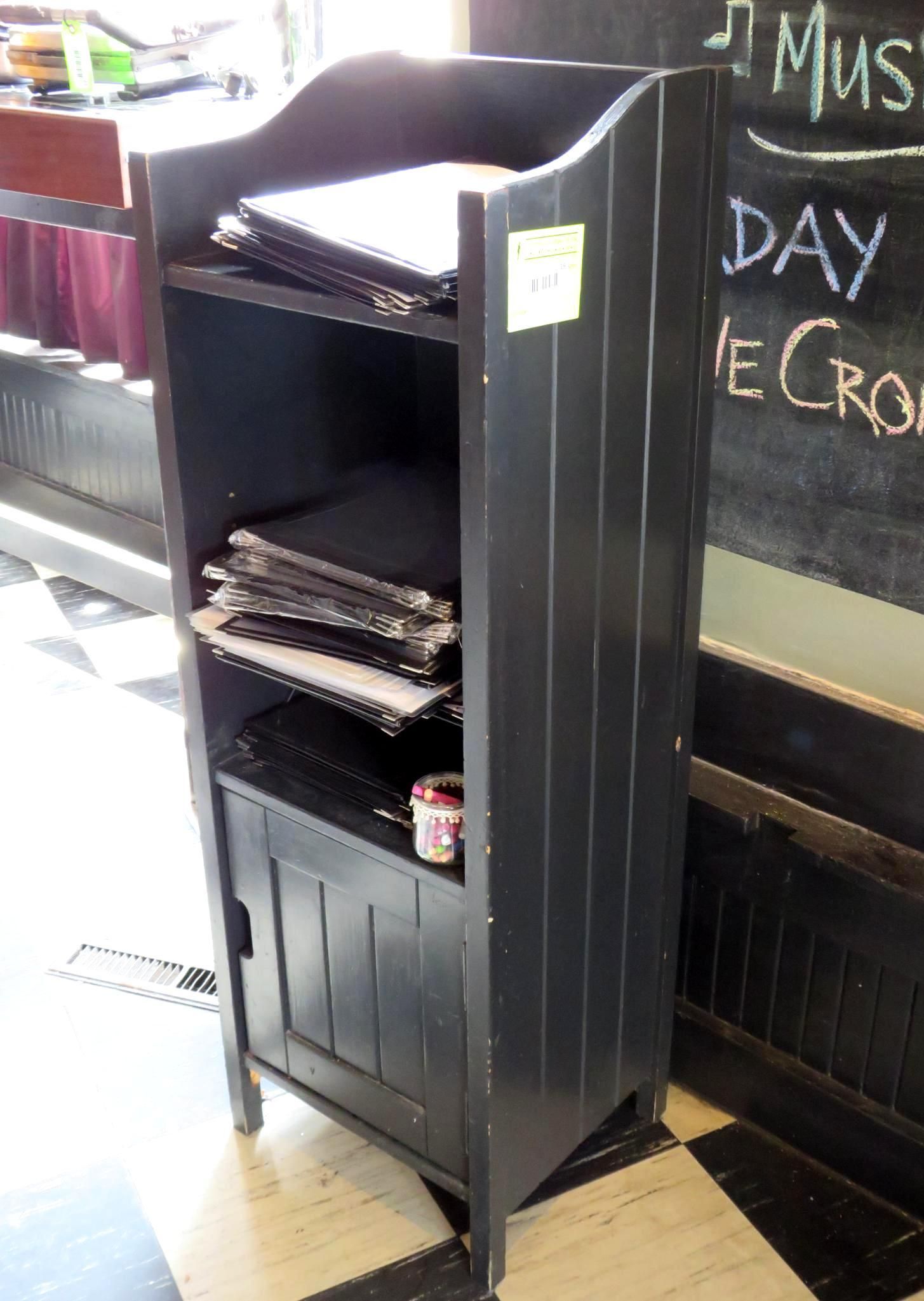 Black menu stand with open shelves and a cabinet, holding menus and a can. Next to a chalkboard.