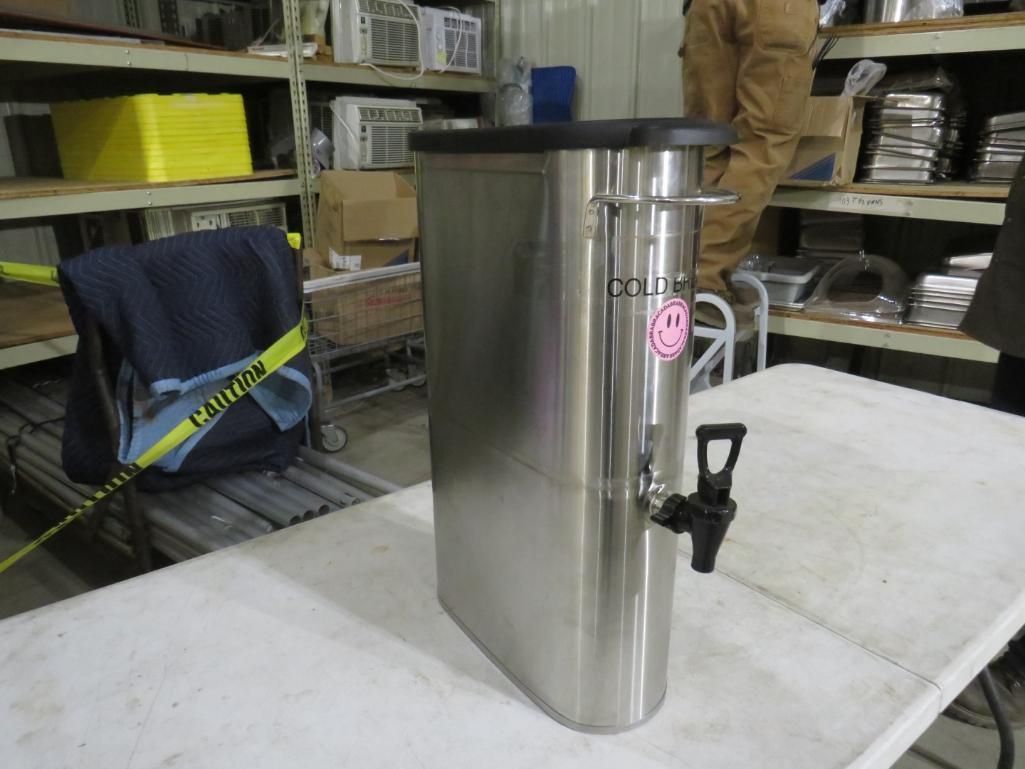 Stainless steel insulated beverage dispenser with a black lid and spigot on a white table.