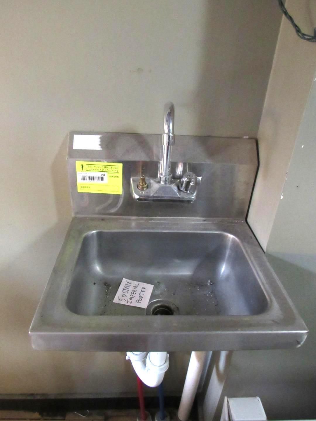 Stainless steel wall-mounted sink with faucet and drain in front of a light gray wall.