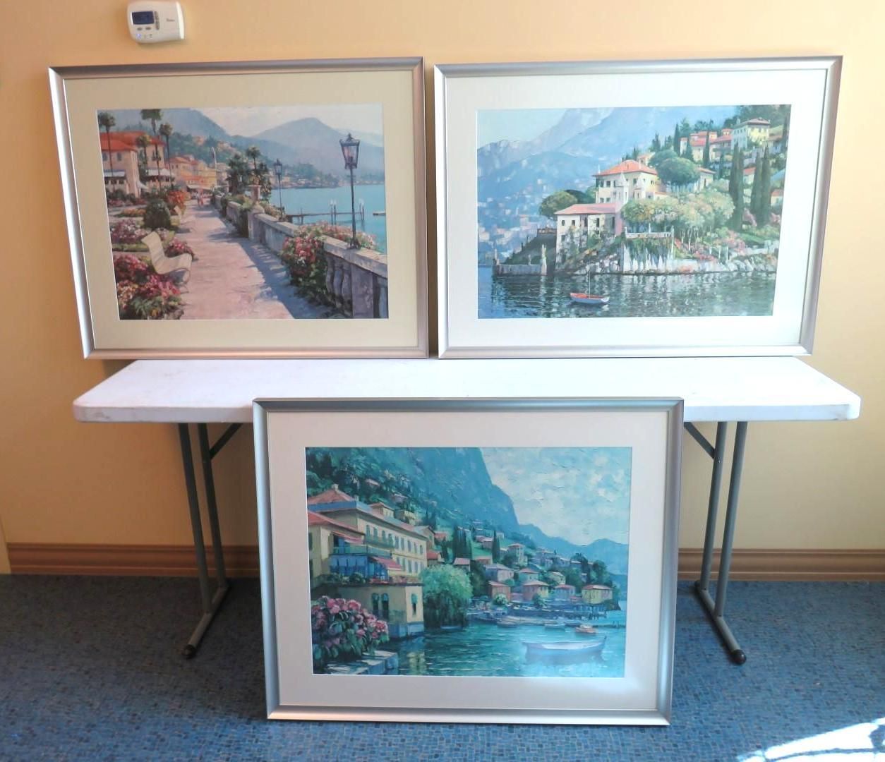 Three framed landscape paintings on a folding table against a yellow wall; a blue carpet.