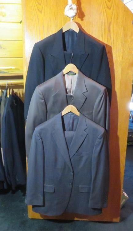Three men's suits in navy, gray, and blue, hanging on a wooden rack.