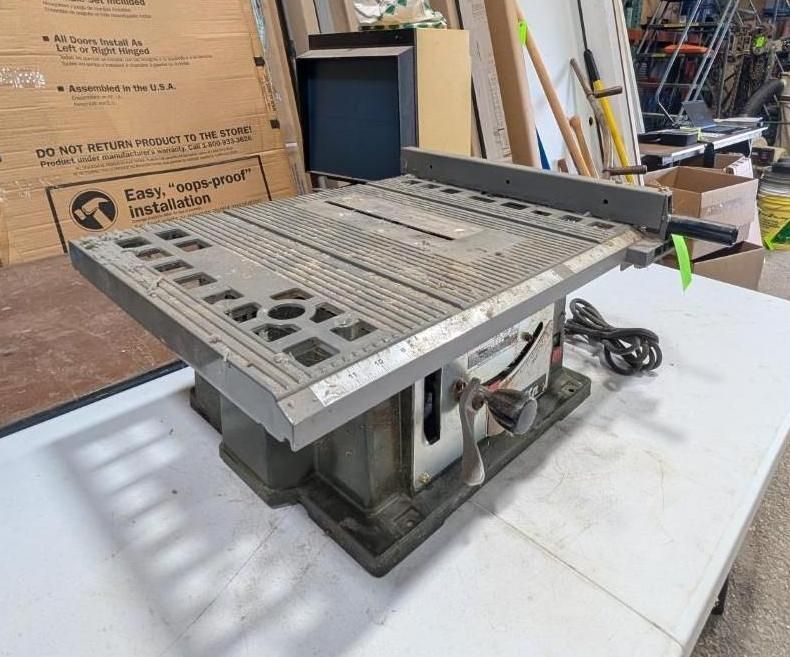 Metal table saw on a workbench in a workshop, with a gray tabletop and angled fence.