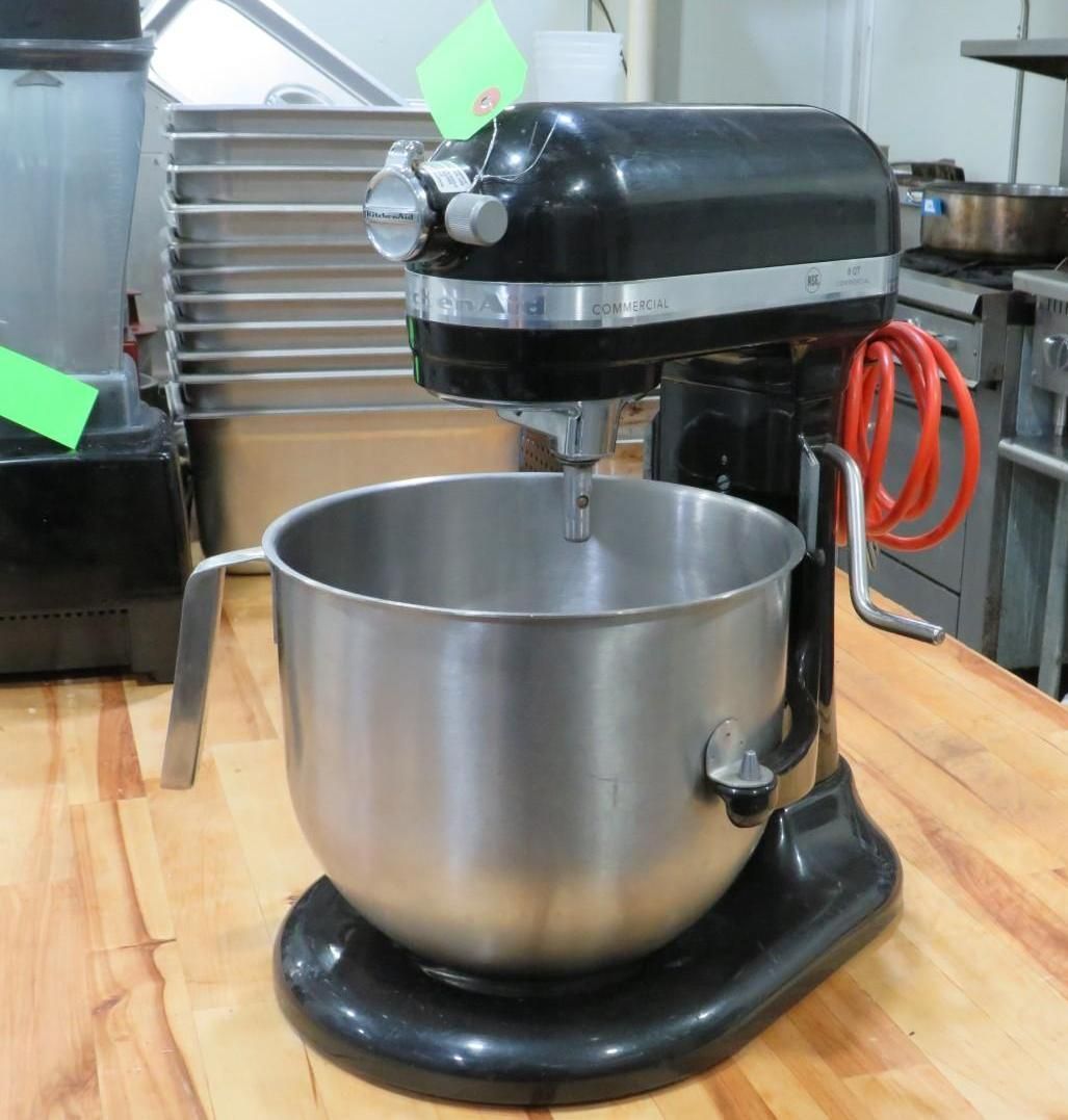 Black stand mixer with stainless steel bowl on a wooden surface, in a commercial kitchen.