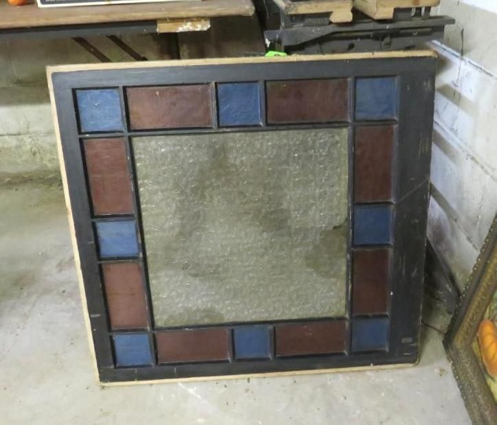 Square stained-glass mirror with blue and brown panes in a black frame, resting on a concrete floor.