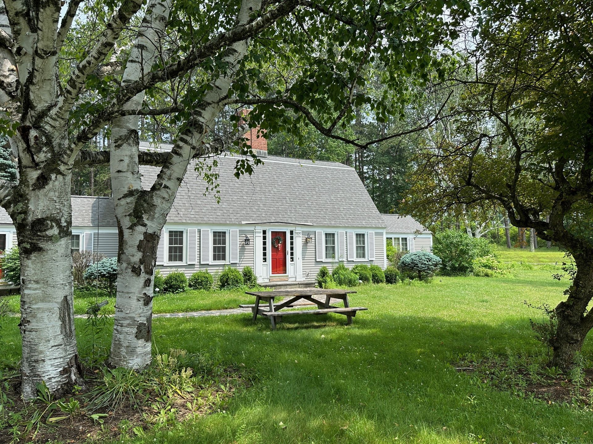 Cottage with a gray roof and red door, flanked by birch trees and a picnic table on the grass.