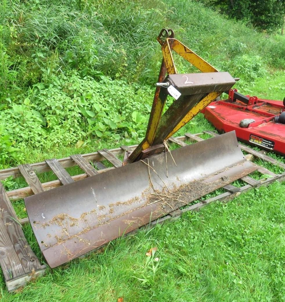 Curved metal blade, yellow frame, attached to a tractor blade, resting on wooden pallet, green grass.