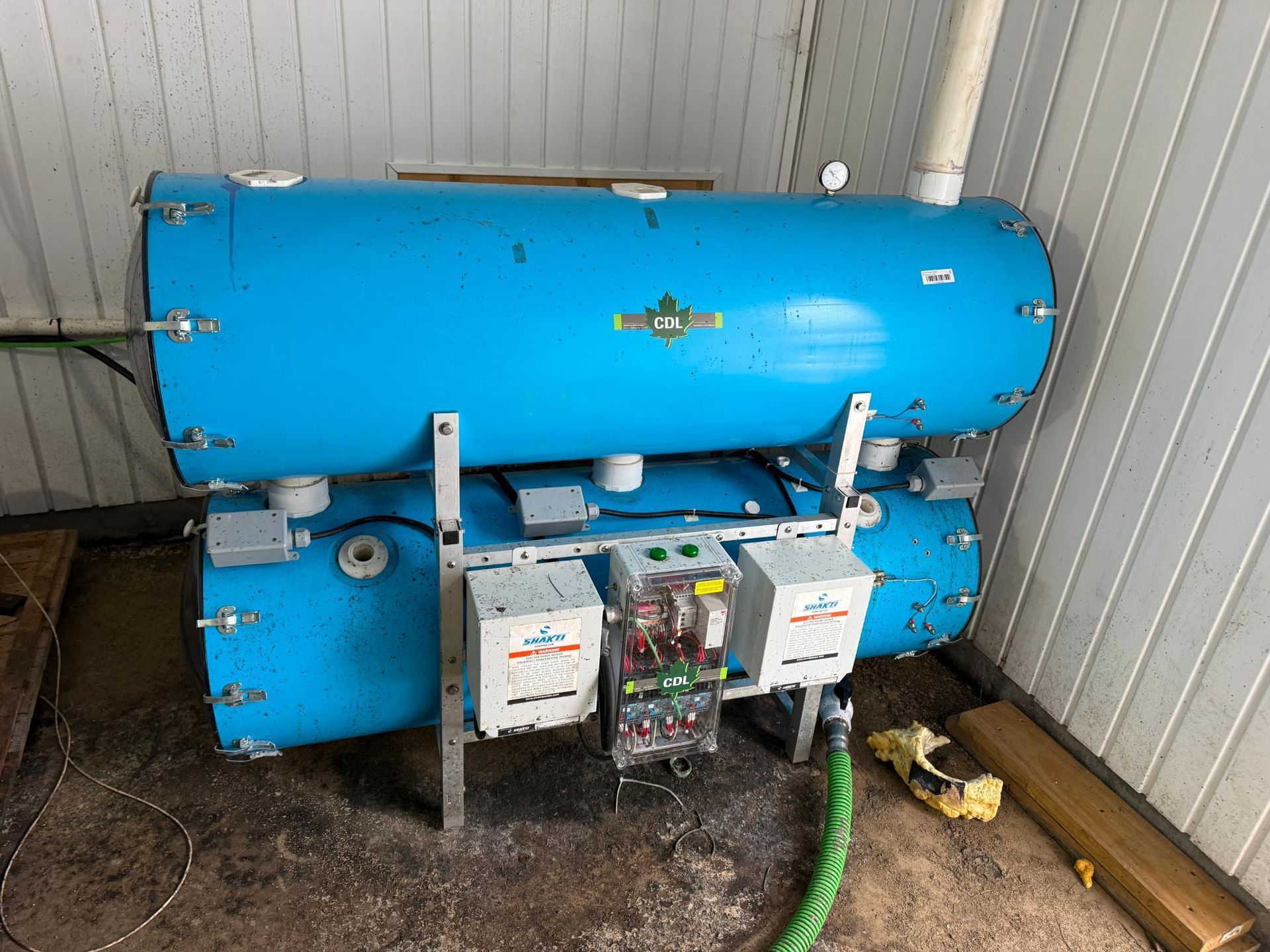 Blue industrial tanks stacked, supported by metal frame, with electrical components and green hose.